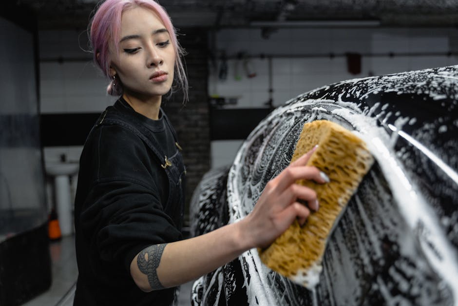 Young woman with pink hair washing a black car using sponge and soap in an indoor carwash setting.